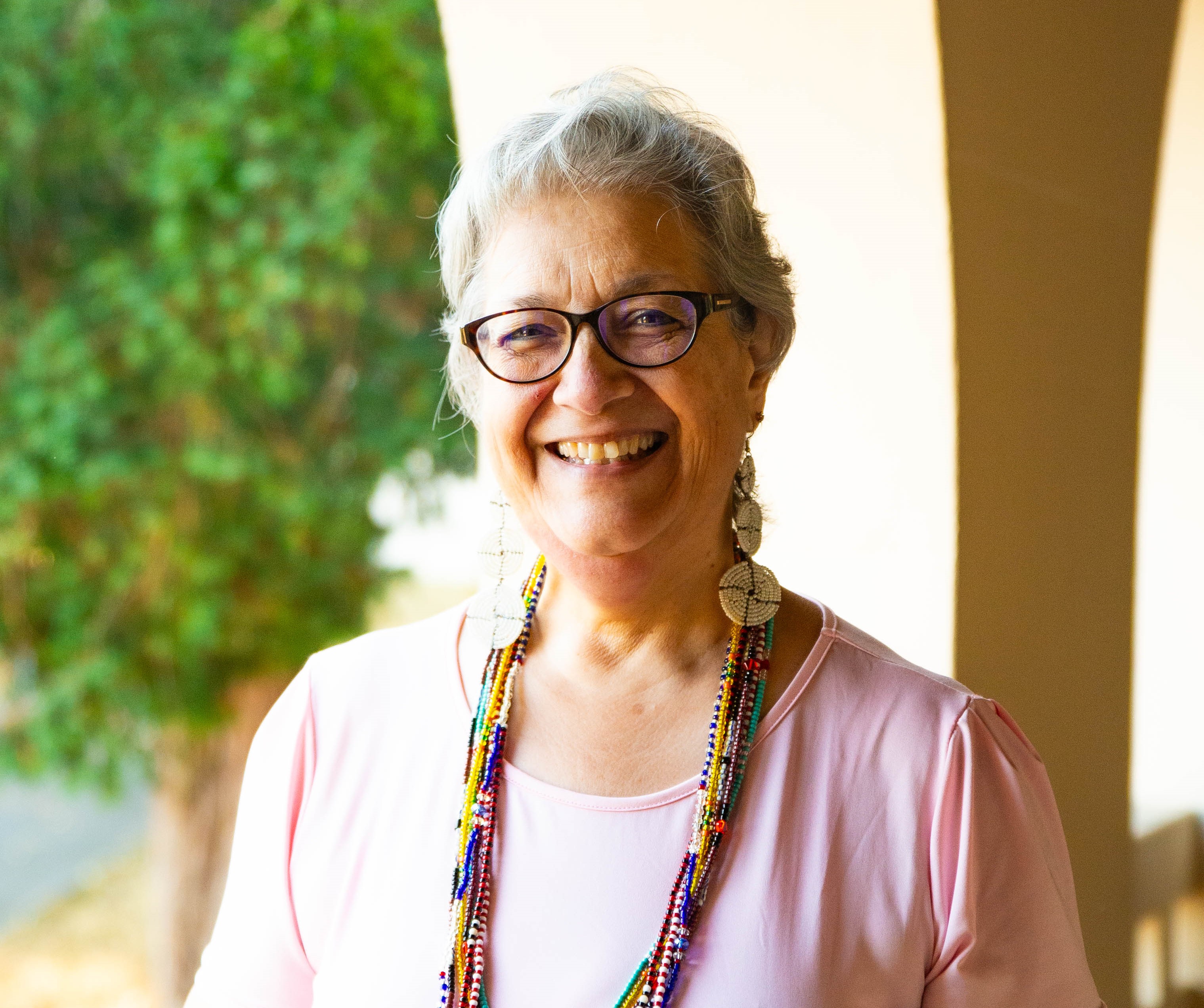 Dr. Louise Santiago standing outside in an arched breezeway with trees behind her. She is wearing a pink shirt, glasses, and beaded necklaces.