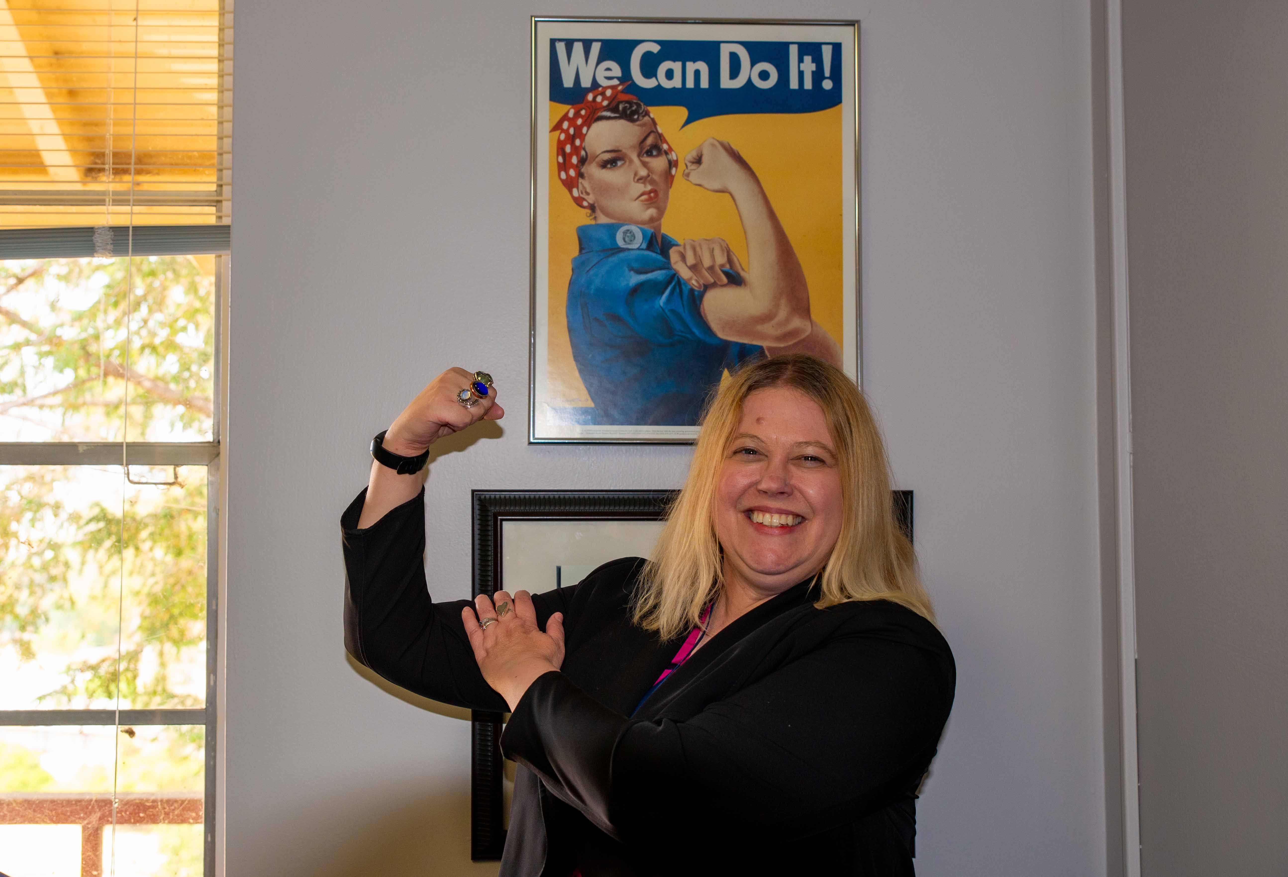 Dr. Lisa Norton Flexes her right arm to flex her bicep underneath a poster of Rosie the riveter in a similar pose with a speech bubble that says, “We can do it!”