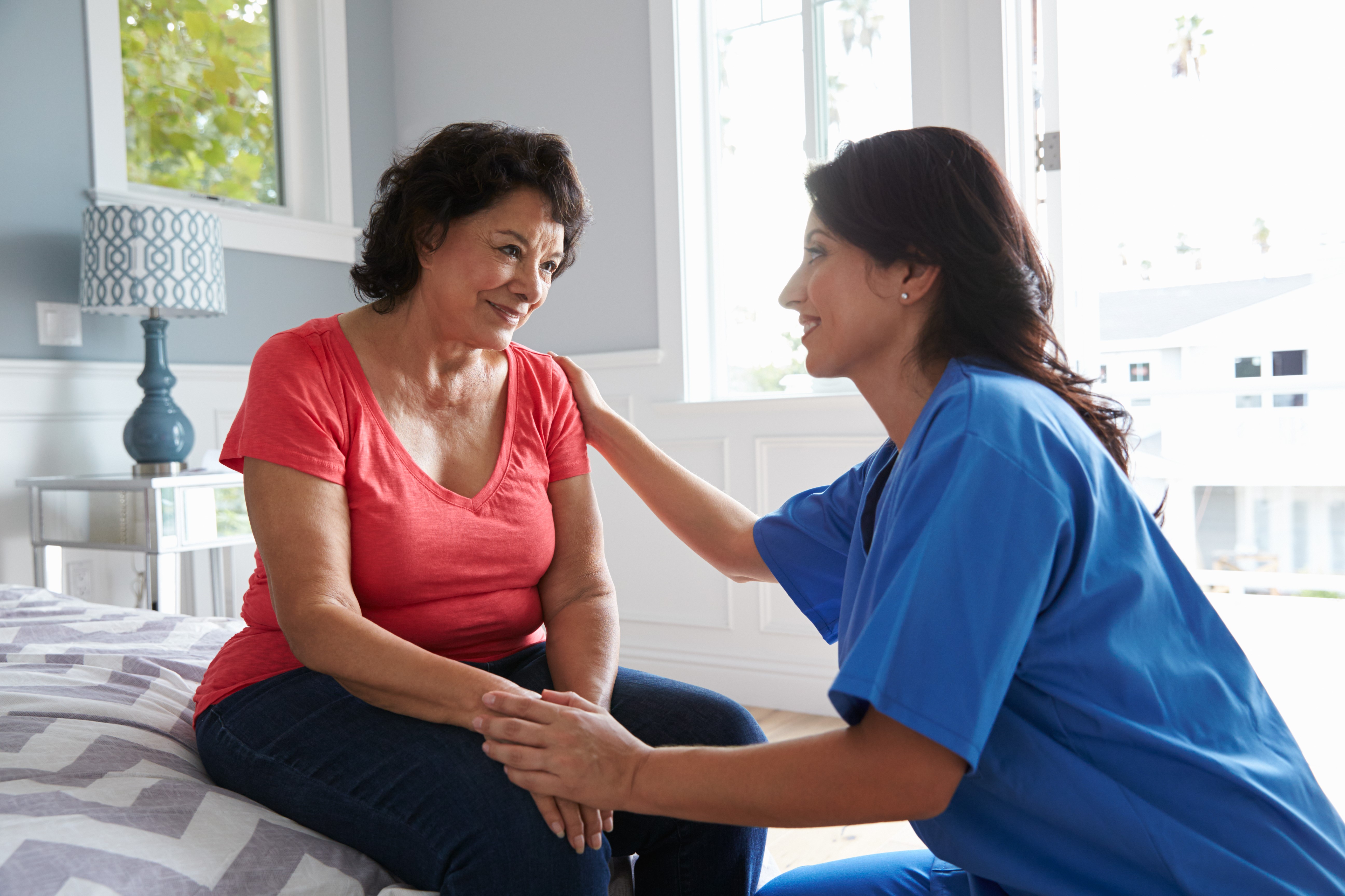 A young nurse in blue scrubs talks to an older woman sitting on the edge of a bed