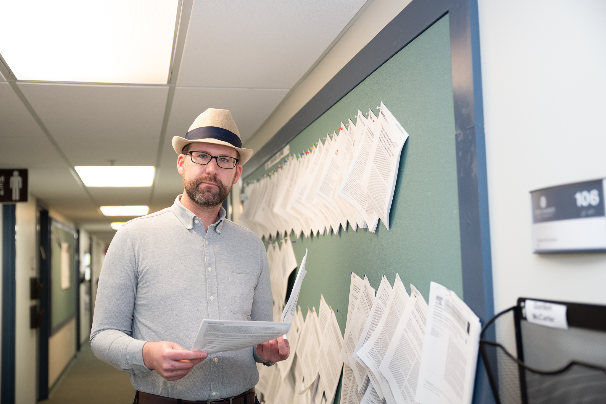 Dr. Kelan Thomas stands in a hall with a cork board lined with research papers, holding one of his studies