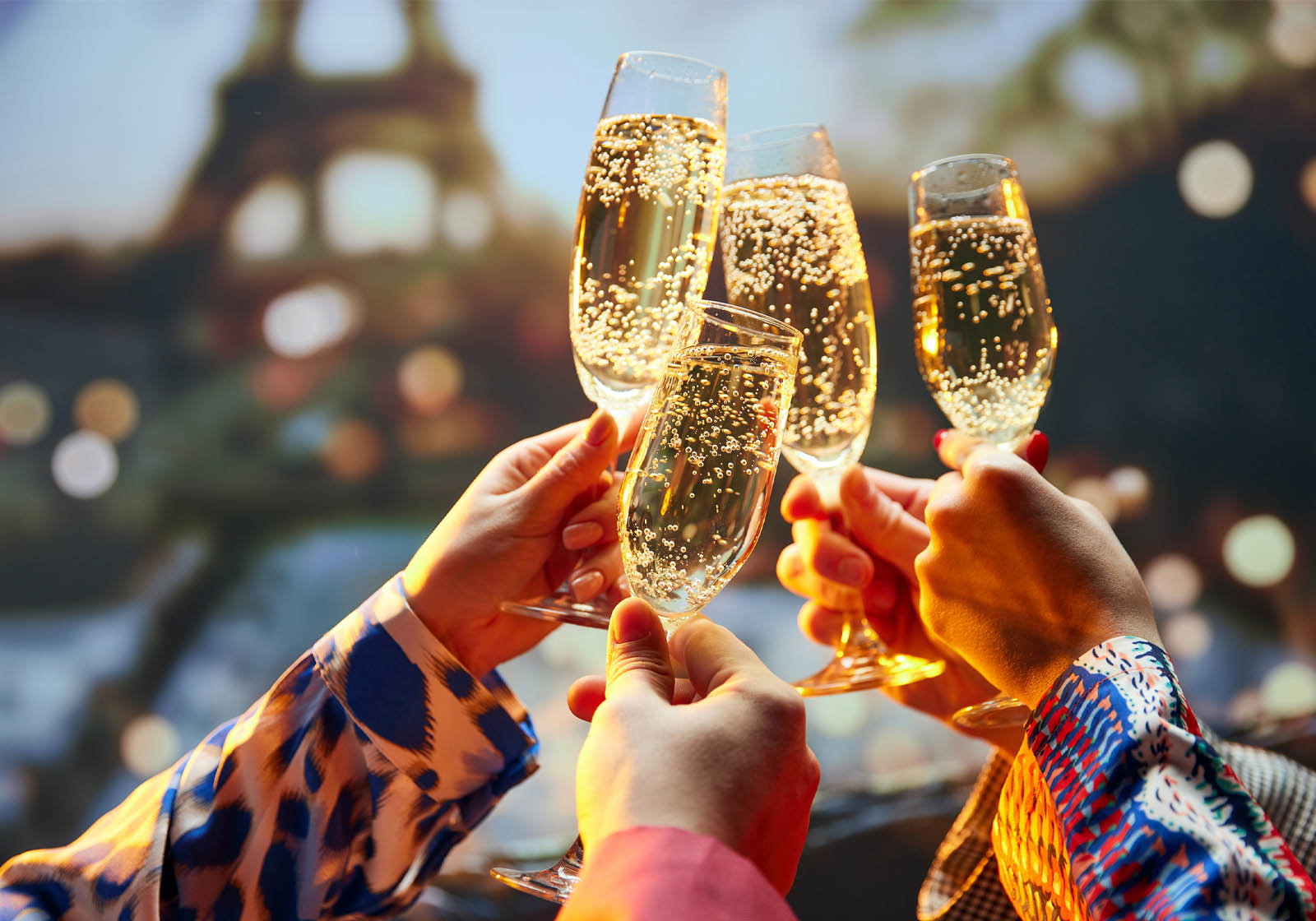 Four hands toasting four full champagne flutes with part of the Eiffel Tower in the background