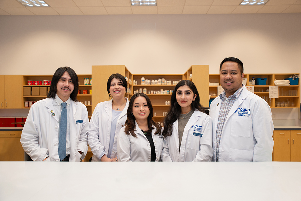 Five Pharmacy students stand together and smile while standing in front of a pharmacy wall