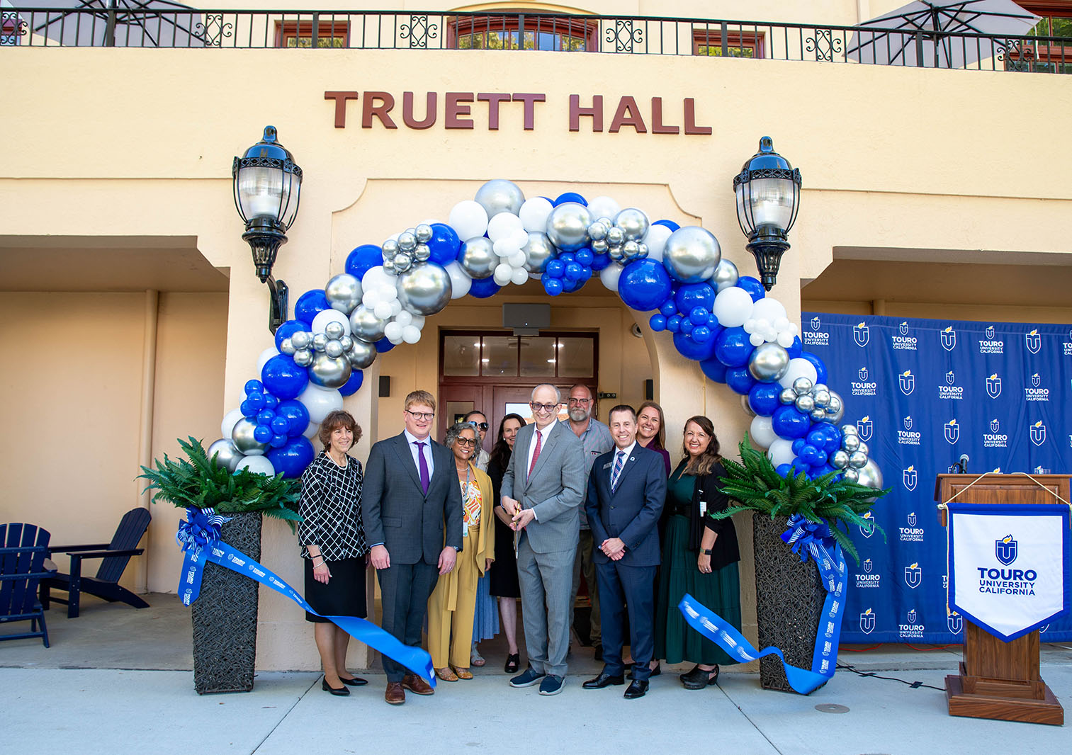 People from Touro University stand in front of the archway of a building and cut a ribbon to celebrate the opening of Truett Hall. 